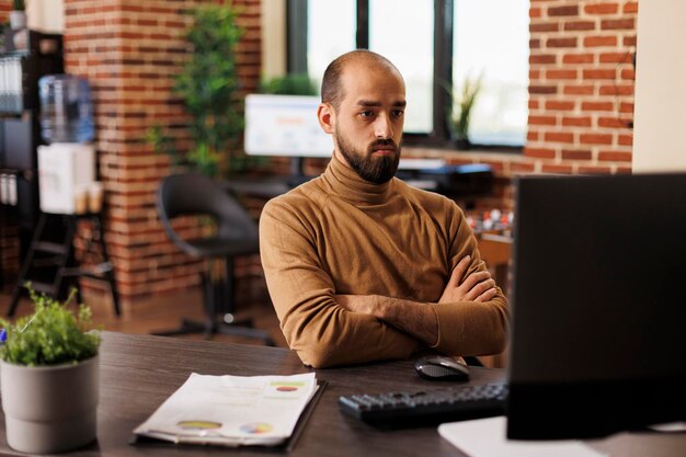 Young businessman sitting in office with crossed arms, looking at computer monitor while reviewing financial statistics. Marketing office employee brainstorming management ideas.