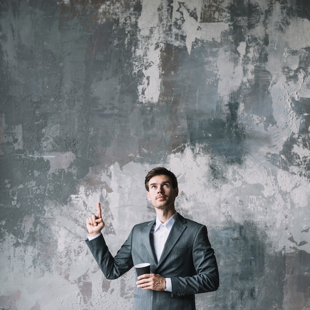Young businessman standing against concrete wall pointing his finger upward looking up