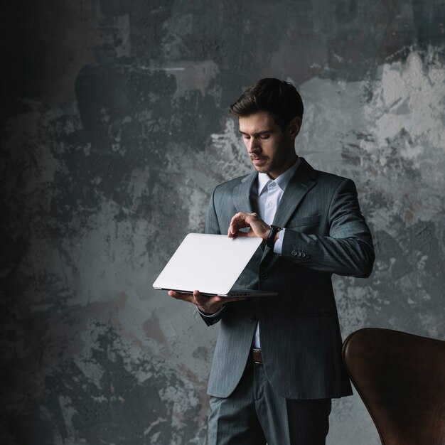 Young businessman standing against an old wall opening the laptop