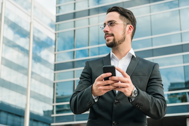 Молодой предприниматель, стоя перед офисное здание, держа мобильный телефон
