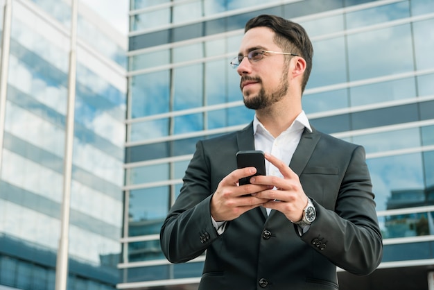 Young businessman standing in front of office building holding mobile phone