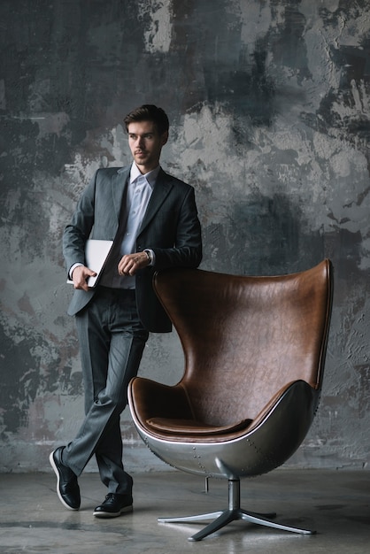 Young businessman standing in front of weathered wall leaning on modern chair holding laptop in hand