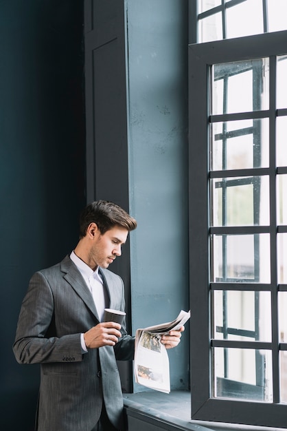 Free photo young businessman standing near the window holding cup of coffee reading newspaper