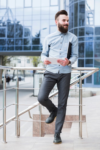 Young businessman standing outside the corporate building holding digital tablet