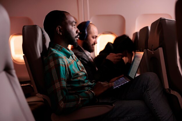 Young businessman travelling in economy class and using laptop during flight, flying abroad on holiday destination or work trip. Working on computer during sunset, aerial transportation.