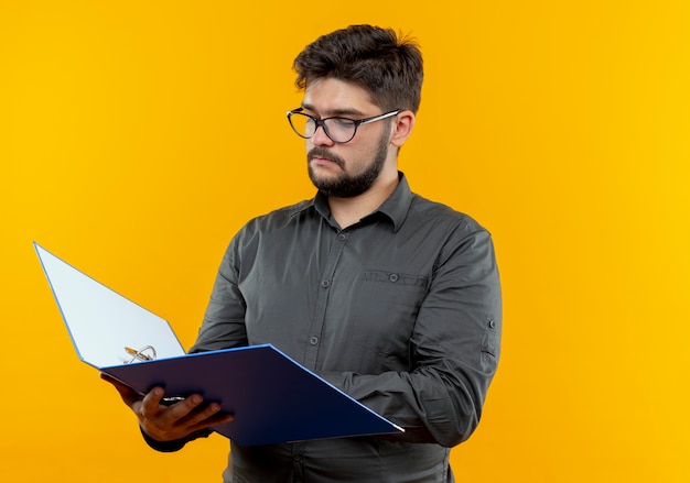 Young businessman wearing glasses holding and looking at folder isolated on yellow