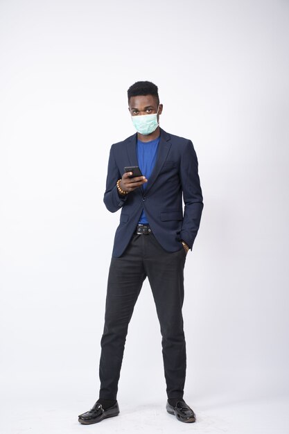 Young businessman wearing a suit and face mask using his phone in front of a white wall