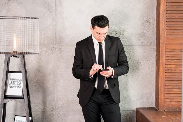 Young businessman with smartphone standing by wall