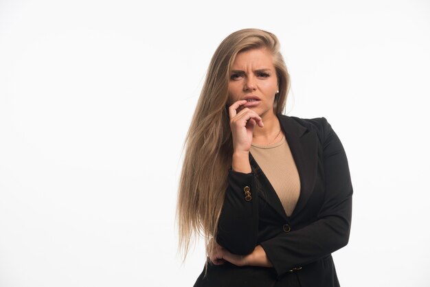 Young businesswoman in black suit looks confused with finger at her mouth.