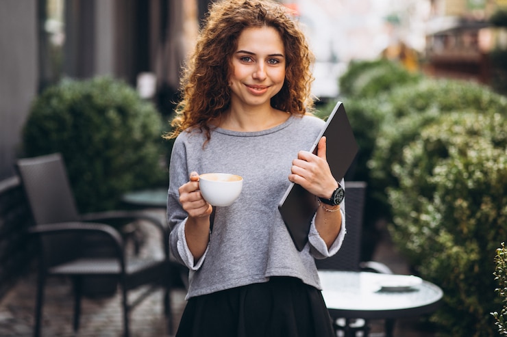 Free Photo young businesswoman drinking coffee outside cafe holding laptop