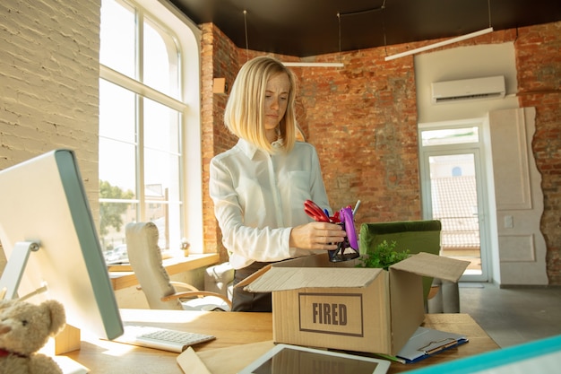 Young businesswoman fired, looks upset. Has to pack her office belongings and to leave work place for new worker. Problems in occupation, stress, unemployment, new way of life or end of career.