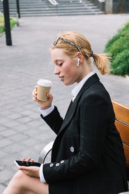 Foto gratuita giovane tazza di caffè della tenuta della donna di affari che esamina telefono cellulare