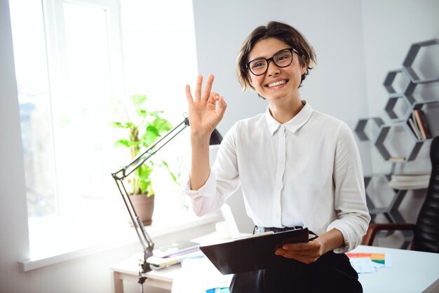 Young businesswoman holding folder, smiling, greeting at workplace in office.