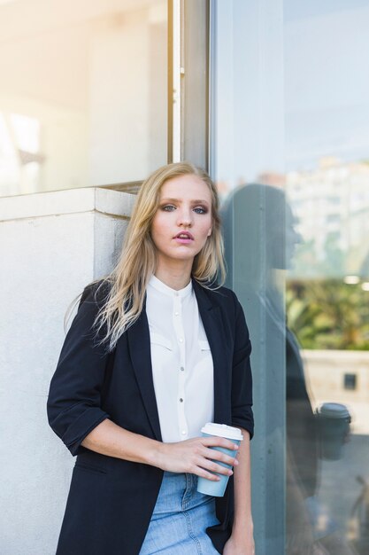 Free photo young businesswoman leaning on wall holding take away coffee cup