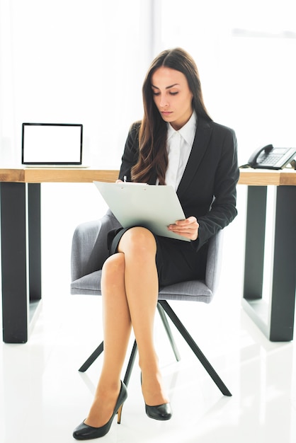 Young businesswoman sitting on chair with crossed legs writing on clipboard