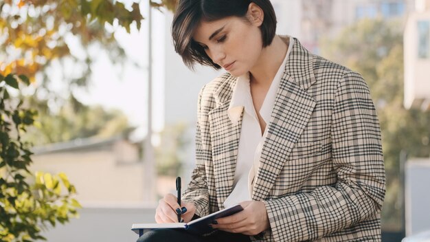 Young businesswoman sitting on street and making notes in her notebook Attractive entrepreneur drawing up business plan for a month outdoor