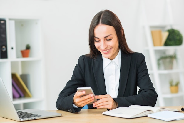 Бесплатное фото Молодая коммерсантка усмехаясь пока смотрящ smartphone в офисе
