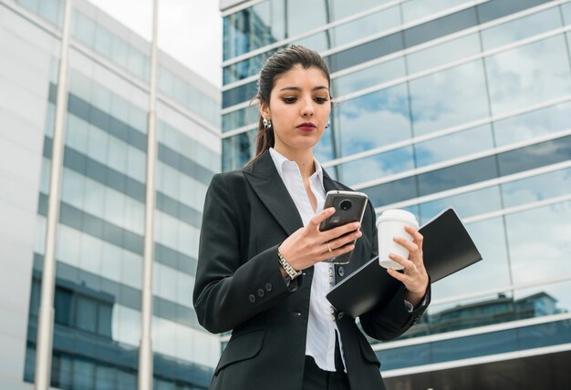 Young businesswoman standing in front of building looking at mobile phone holding takeaway coffee cup in hand