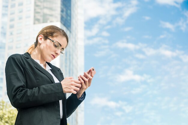 Молодая коммерсантка стоя перед сообщением здания отправляя СМС на мобильном телефоне