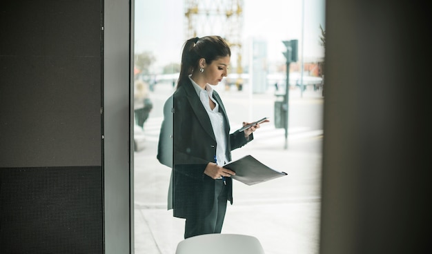 Young businesswoman standing outside the glass door using mobile phone