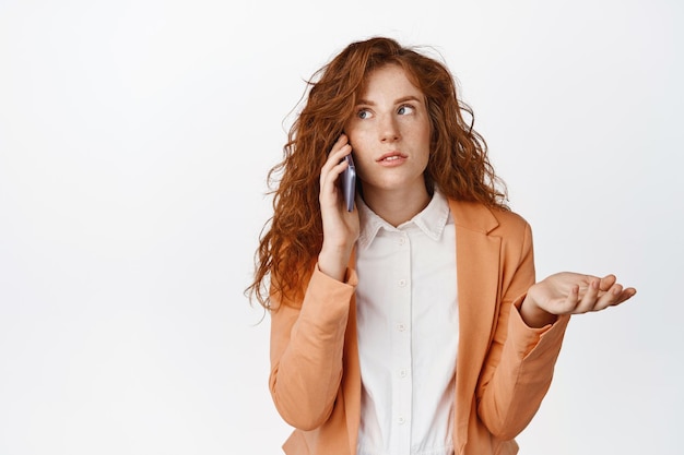 Foto gratuita giovane imprenditrice parlando al cellulare e guardando confuso avendo telefonata aziendale in piedi in tuta su sfondo bianco