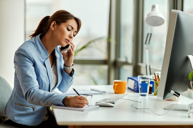 Young businesswoman talking on mobile phone while writing notes and working in the office