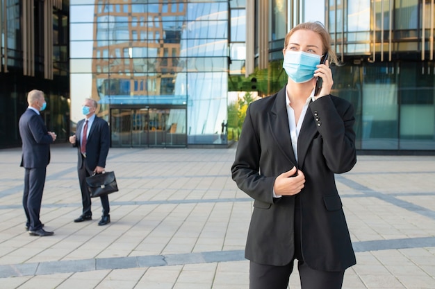 Free photo young businesswoman wearing mask and office suit talking on cellphone outdoors. businesspeople and city buildings in background. copy space. business and epidemic concept