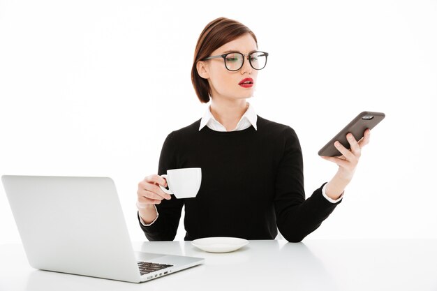Young businesswoman with coffee or tea cup and laptop computer, using smartphone