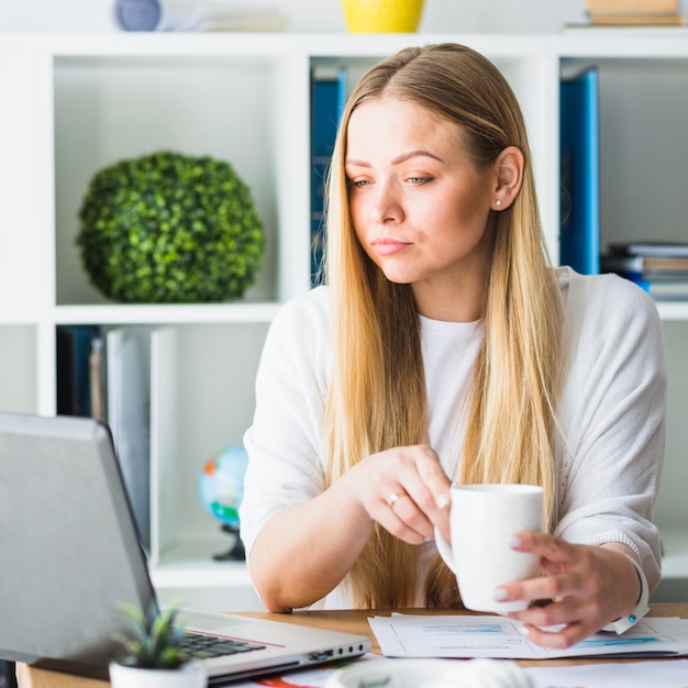 Free photo young businesswoman with cup of coffee looking at laptop