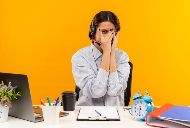 Young call center girl wearing headset sitting at desk with work tools covering face with hands looking at camera through fingers isolated on orange background