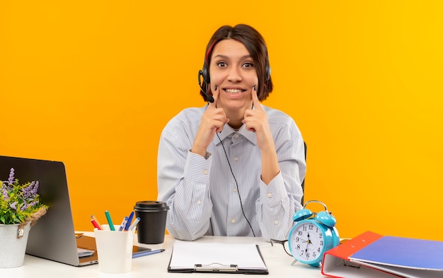 Foto gratuita giovane ragazza del call center che indossa l'auricolare seduto alla scrivania con strumenti di lavoro fingendo sorriso guardando la telecamera isolata su sfondo arancione