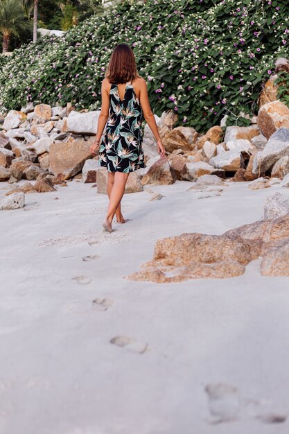 Young calm tattooed woman in summer tropical print short dress on rocky beach with green bush and purple pink flowers