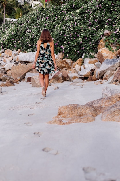 Free photo young calm tattooed woman in summer tropical print short dress on rocky beach with green bush and purple pink flowers