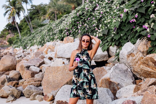 Young calm tattooed woman in summer tropical print short dress on rocky beach with green bush and purple pink flowers