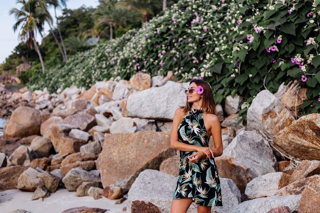 Young calm tattooed woman in summer tropical print short dress on rocky beach with green bush and purple pink flowers