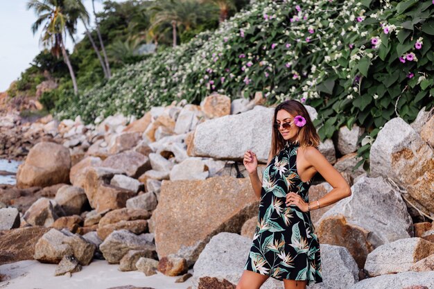 Young calm tattooed woman in summer tropical print short dress on rocky beach with green bush and purple pink flowers