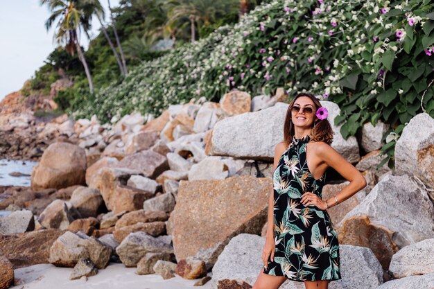 Young calm tattooed woman in summer tropical print short dress on rocky beach with green bush and purple pink flowers