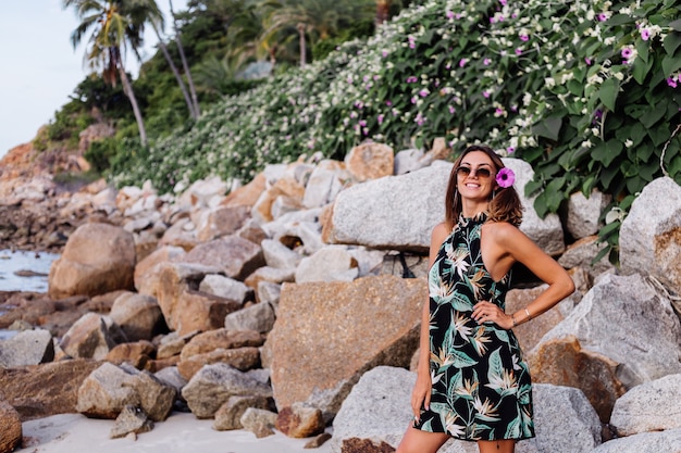 Young calm tattooed woman in summer tropical print short dress on rocky beach with green bush and purple pink flowers