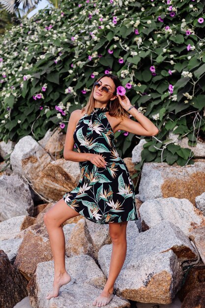 緑の茂みと紫のピンクの花と岩のビーチで夏の熱帯プリントショートドレスの若い穏やかな入れ墨の女性