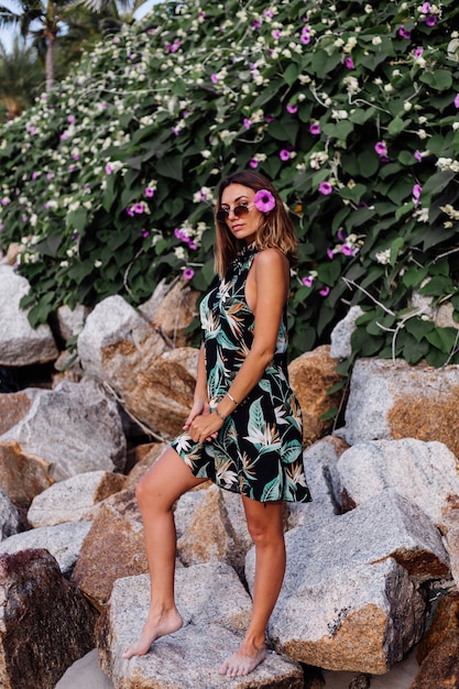 緑の茂みと紫のピンクの花と岩のビーチで夏の熱帯プリントショートドレスの若い穏やかな入れ墨の女性