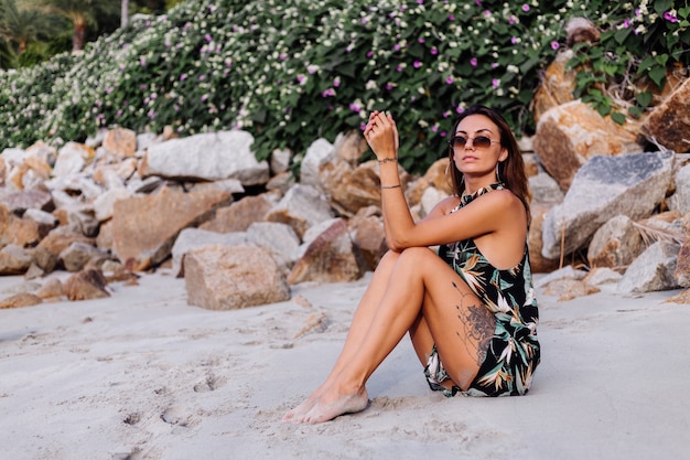 Young calm tattooed woman in summer tropical print short dress on rocky beach with green bush and purple pink flowers