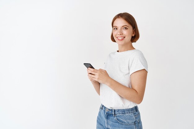 Young candid girl using smartphone and smiling . Woman chatting on mobile phone app, standing against white wall