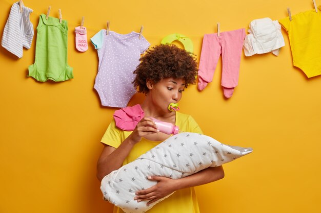 Young caring mother holds infant son wrapped in duvet on hands, feeds with milk from bottle, busy with motherhood, poses at home with washed baby clothes hanging in wall. Family concept