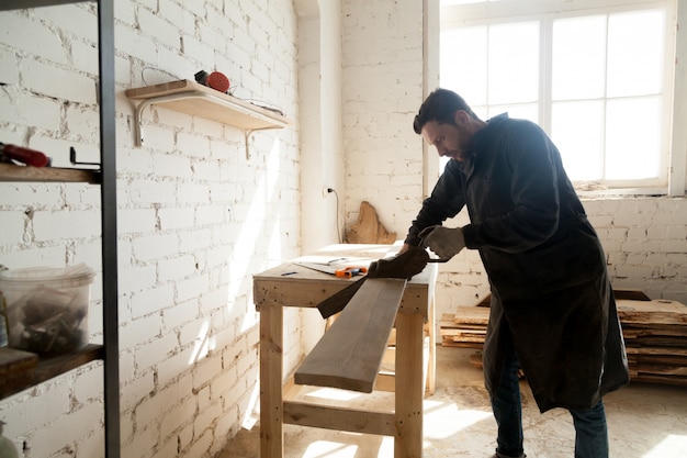 Young carpenter cutting wooden board with hand saw in workshop