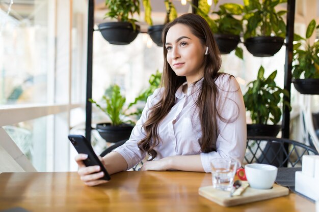 Young casual businesswoman drinking coffee while using phone and listening music on earphones in a cafe.