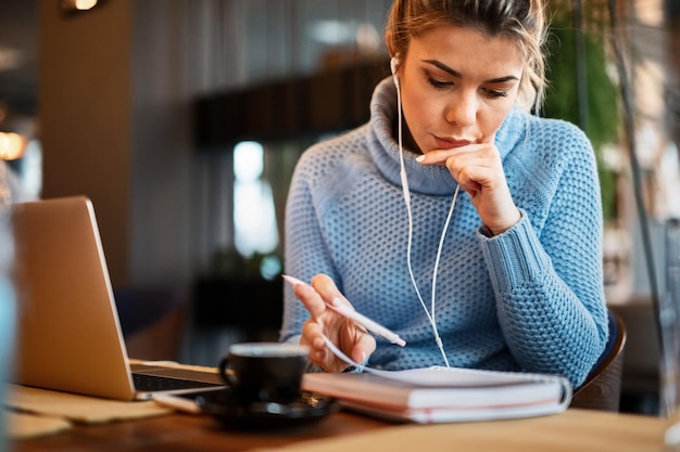 Young casual businesswoman working in a cafe and going though paperwork while listening music over earphones