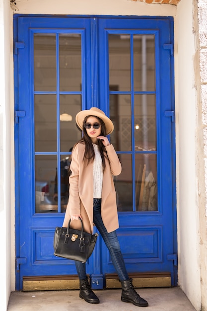 明るいコートの帽子とサングラスの若いカジュアルな服を着た女性は青いドアの前に立っています。