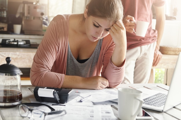 Young casual female having depressed look while managing family finances and doing paperwork, sitting at kitchen table with lots of papers, calculator and laptop