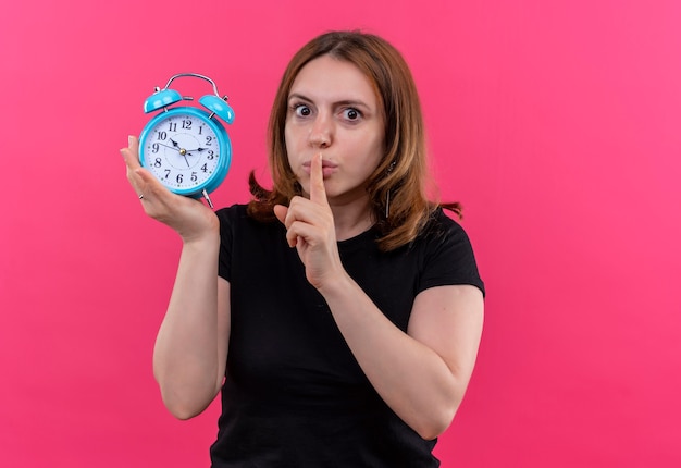 Young casual woman holding alarm clock and gesturing silence on isolated pink wall with copy space