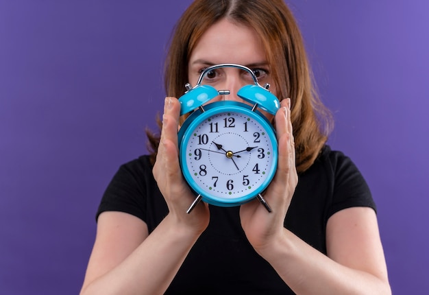 Young casual woman holding and hiding behind alarm clock and looking at it on isolated purple wall